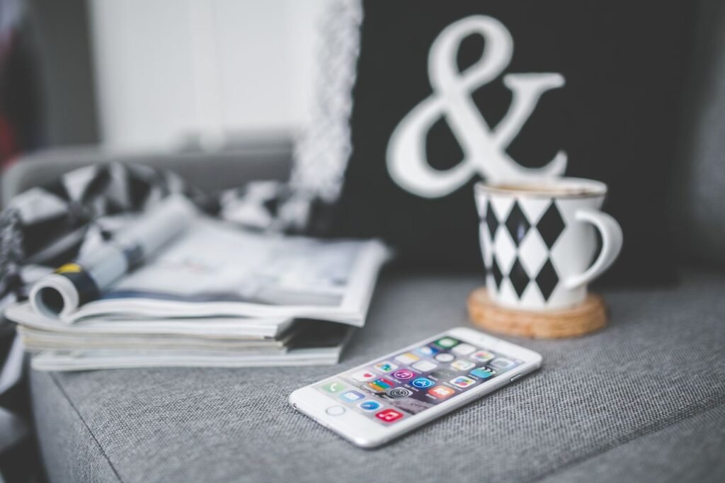A smartphone next to a stylish coffee cup and magazines on a cozy couch setting.